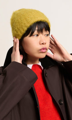 Plümo  Person in an Avo Wool Beanie, red sweater, and brown coat looks to the side with hands by their ears.
