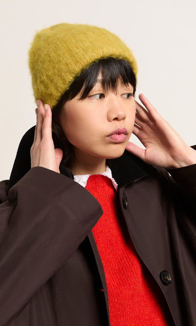 Plümo  Person in an Avo Wool Beanie, red sweater, and brown coat looks to the side with hands by their ears.