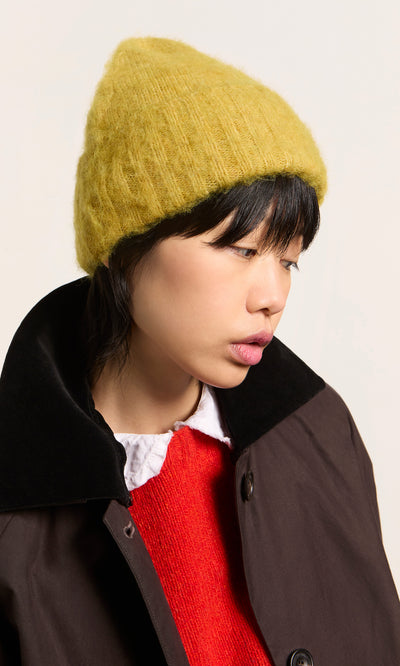 Plümo  Person in an Avo Wool Beanie, red sweater, and brown coat looks downward against a plain background.