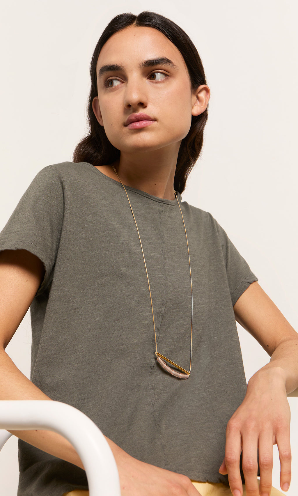 Plümo  Person wearing a Grey Cotton Tee and a long gold necklace, sitting sideways against a plain background.
