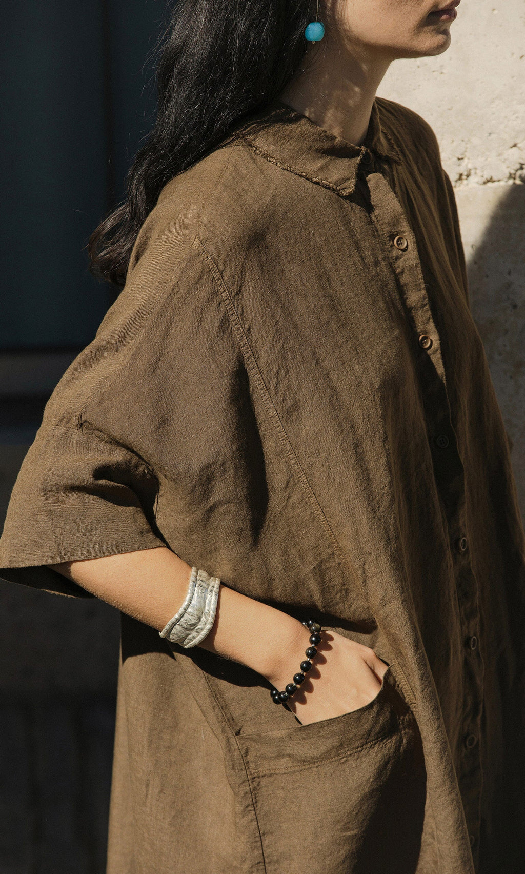 Plümo- Woman in a loose brown button-up dress, black beaded bracelet, and the Bastion Bangle in recycled silver.