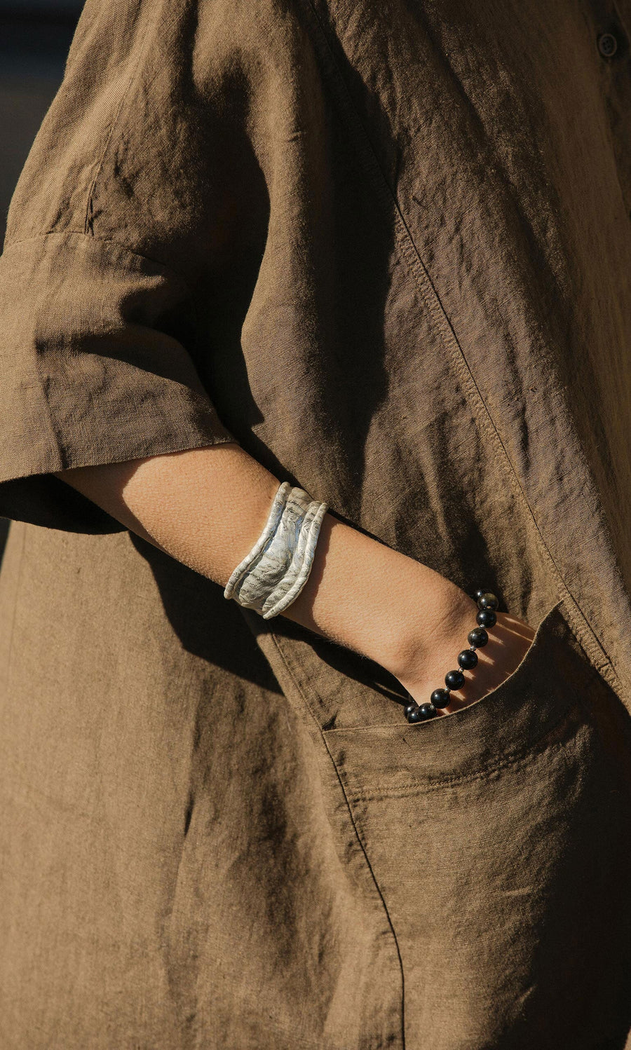 Plümo- Someone in a brown shirt wears the Bastion Bangle, an organically shaped accessory, with a hand in their pocket.