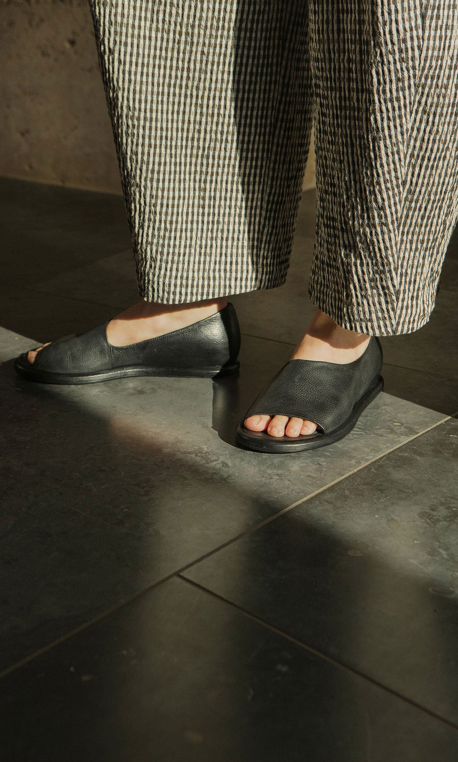 Plümo- Person wearing Hawke Sandals and checkered pants stands on a sunlit tiled floor.