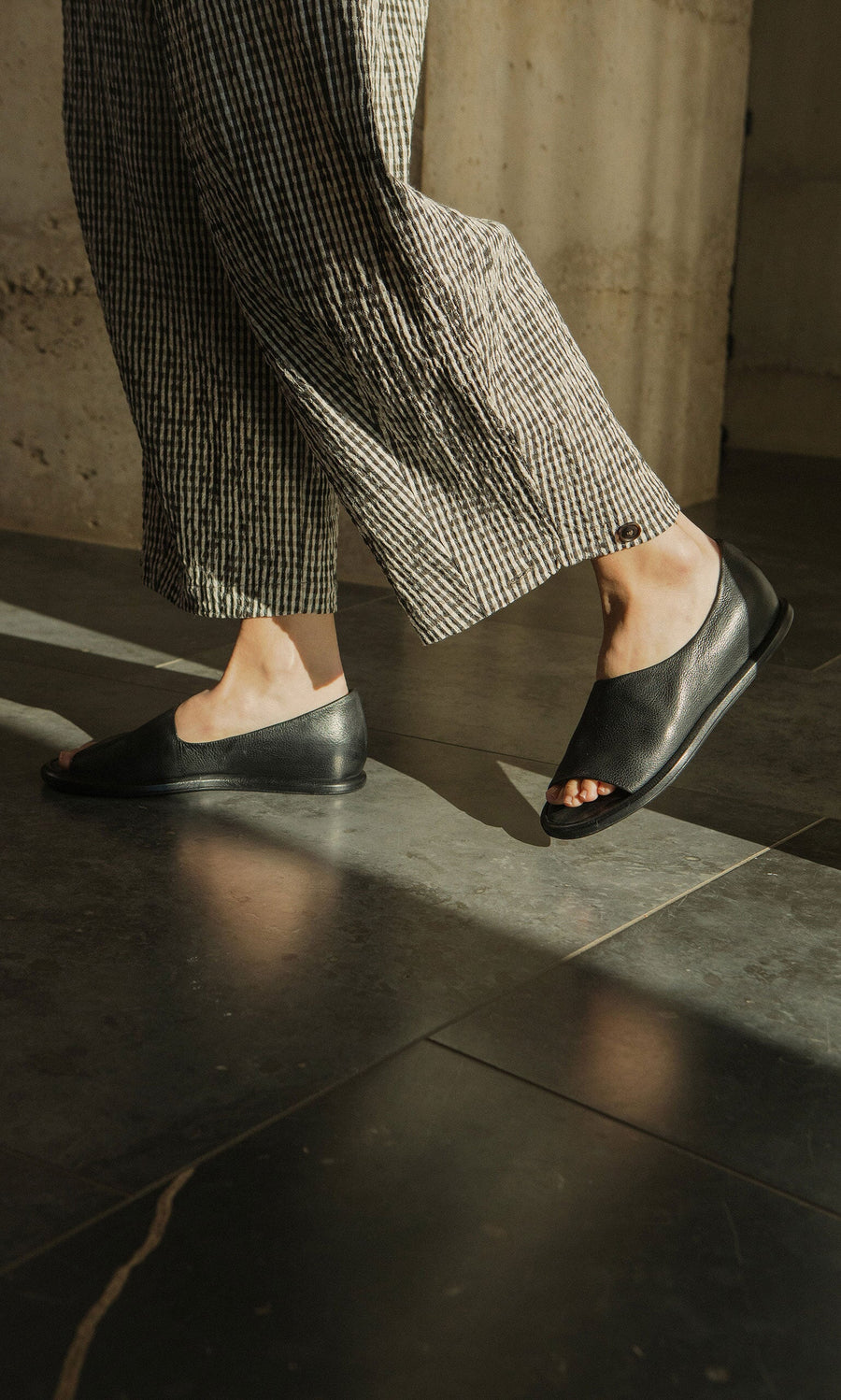 Plümo- Person wearing Hawke Sandals and striped pants walks on a sunlit tiled floor.