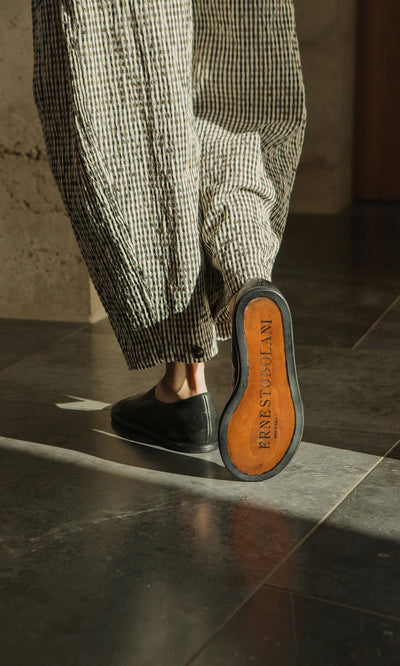 Plümo- Person in checkered pants wearing Hawke Sandals; sole reads FARFETCH X COLLAN as they walk on a tiled floor.