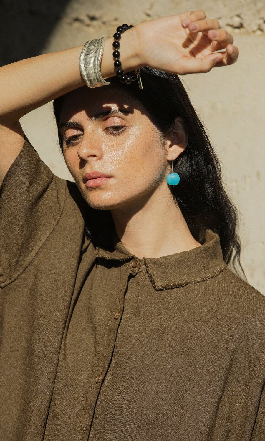 Plümo- Woman in a brown collared shirt wears Bola Earrings and handmade jewellery, shielding her eyes from sunlight.