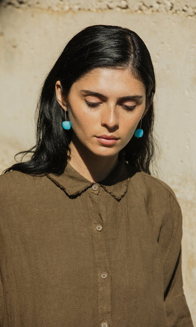 Plümo- Woman with long dark hair in a brown shirt wears Bola Earrings, standing outside with eyes closed in sunlight.