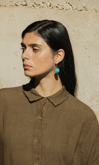 Plümo- Woman with long dark hair in a brown shirt wears Bola Earrings in cerulean blue, standing by a beige wall.
