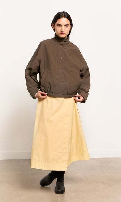 Plümo- Person wearing a Bomber Jacket, light yellow skirt, and black boots against a plain background.