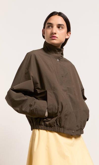 Plümo- Someone in an olive-green Danish design Bomber Jacket and yellow skirt looks sideways against a plain background.