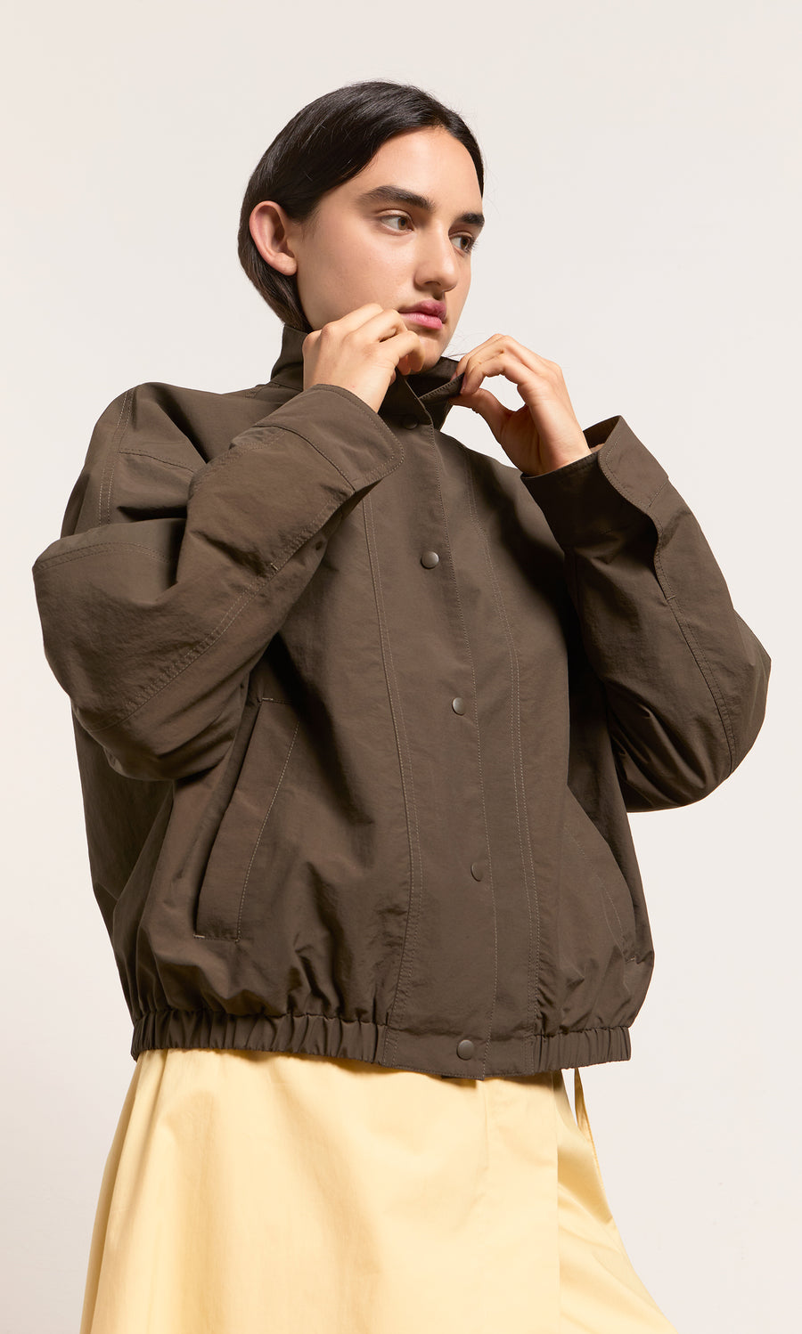 Plümo- Person in a loose Danish design Bomber Jacket over a light yellow dress, adjusting the collar against a plain backdrop.