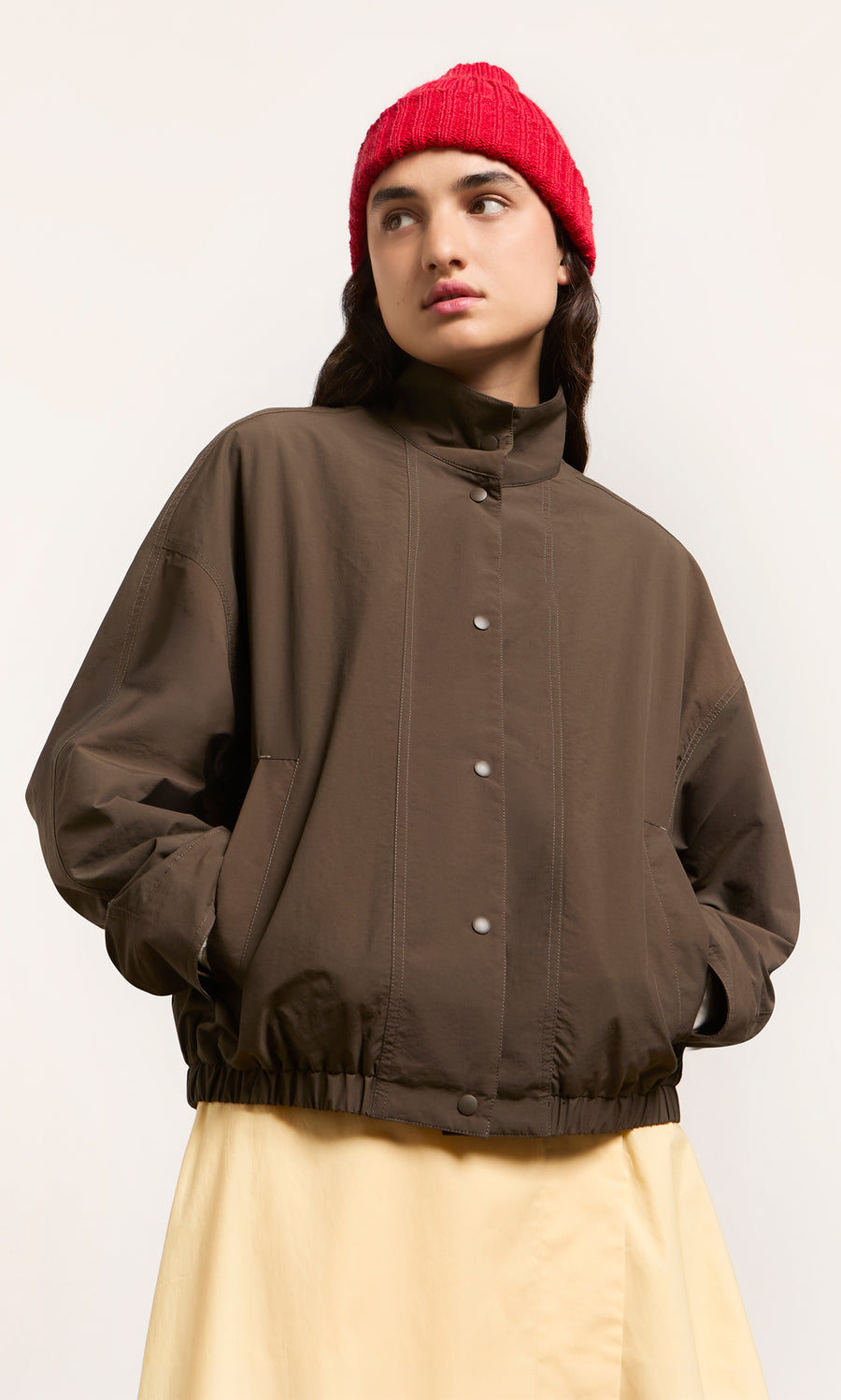 Plümo  Individual in a brown jacket, Red Breton Beanie, and yellow skirt stands against a plain light background.