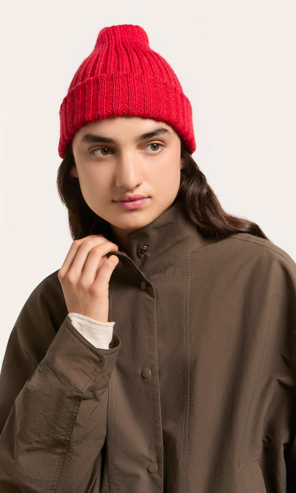 Plümo  Individual wearing a Red Breton Beanie and olive jacket gazes sideways against a simple backdrop.