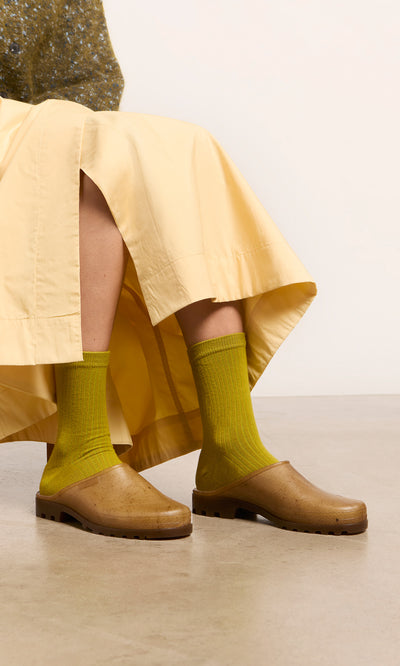 Plümo  Person in a yellow skirt, bright green socks, and brown clogs sits on a chair against a plain background.