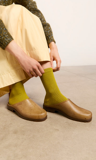 Plümo  Person in bright green socks and tan clogs adjusts pants while sitting on a concrete floor.