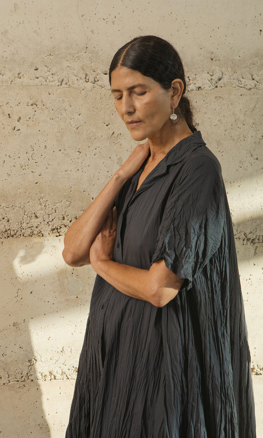 Plümo- Woman in a crinkled dark dress stands by a textured wall, eyes closed, wearing Shirley Earrings.