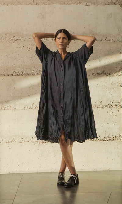Plümo- Woman wears the Brook Crinkle Dress, standing with arms raised against a textured beige wall.