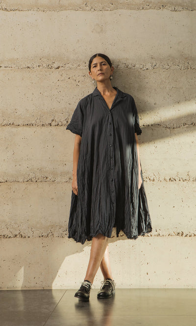 Plümo- Woman in the Brook Crinkle Dress stands against a textured beige wall, looking upwards.