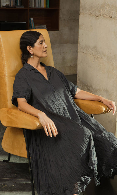 Plümo- Woman in the Brook Crinkle Dress relaxes in a tan armchair, eyes closed, against a concrete wall.