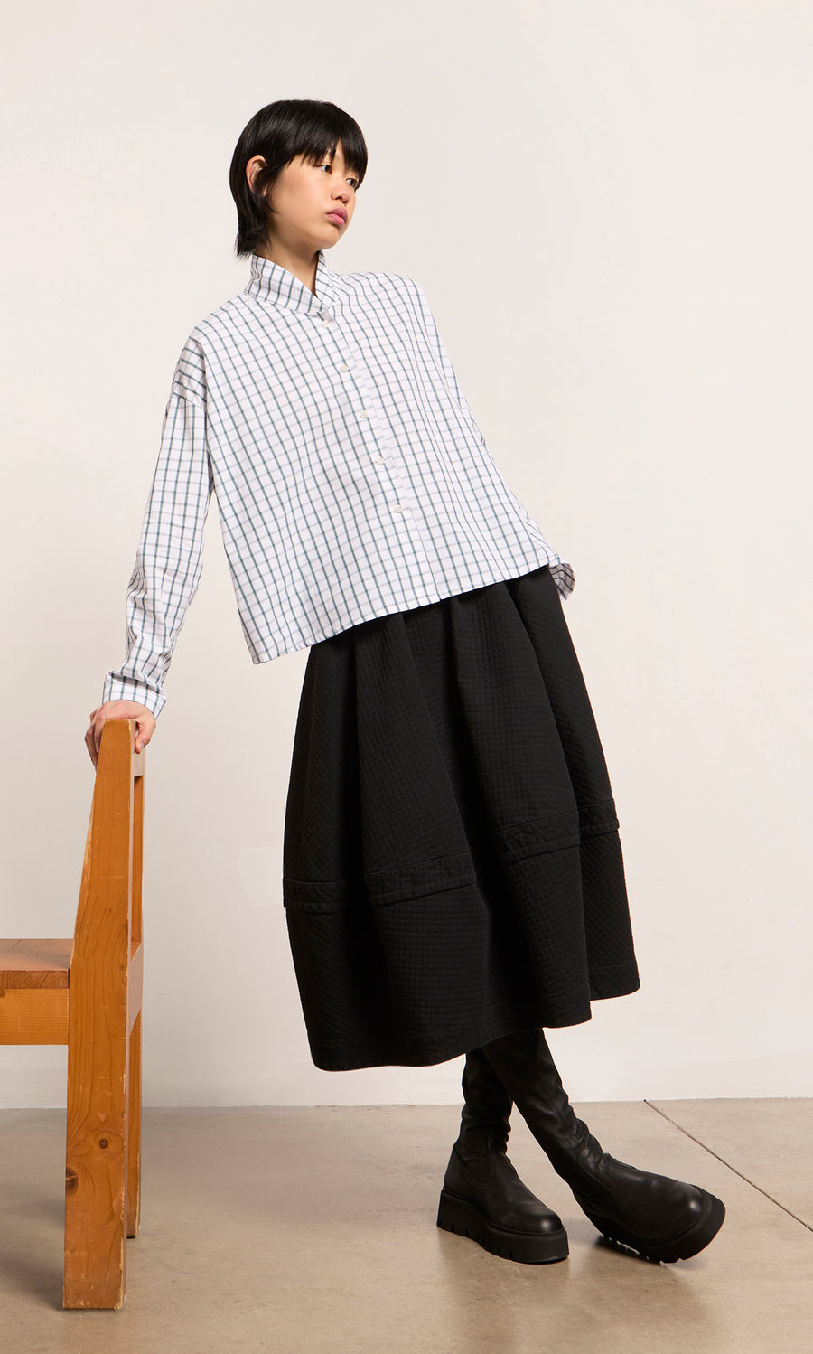 Plümo  Person in a Bruges Shirt, black skirt, and boots leans on a wooden chair against a plain background.