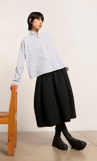 Plümo  Person in a checkered shirt, Rho Skirt - Coal, and boots leans on a wooden chair in a minimalist room.