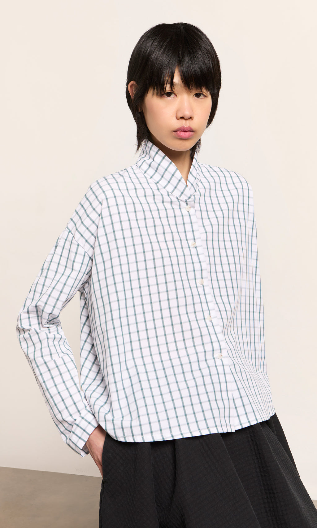 Plümo  Person in the green grid check Bruges Shirt and black pants stands hands-in-pockets against a plain background.