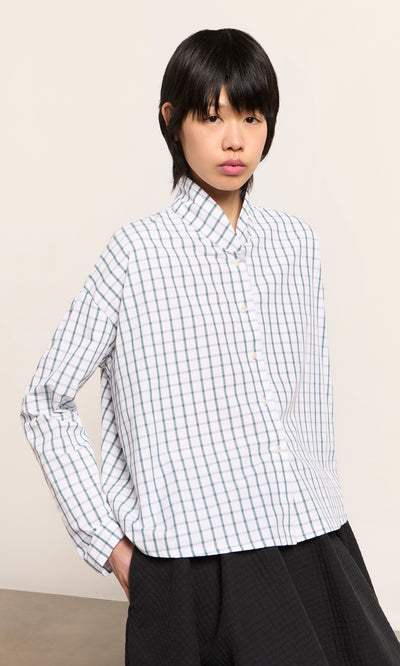 Plümo  Person in the green grid check Bruges Shirt and black pants stands hands-in-pockets against a plain background.