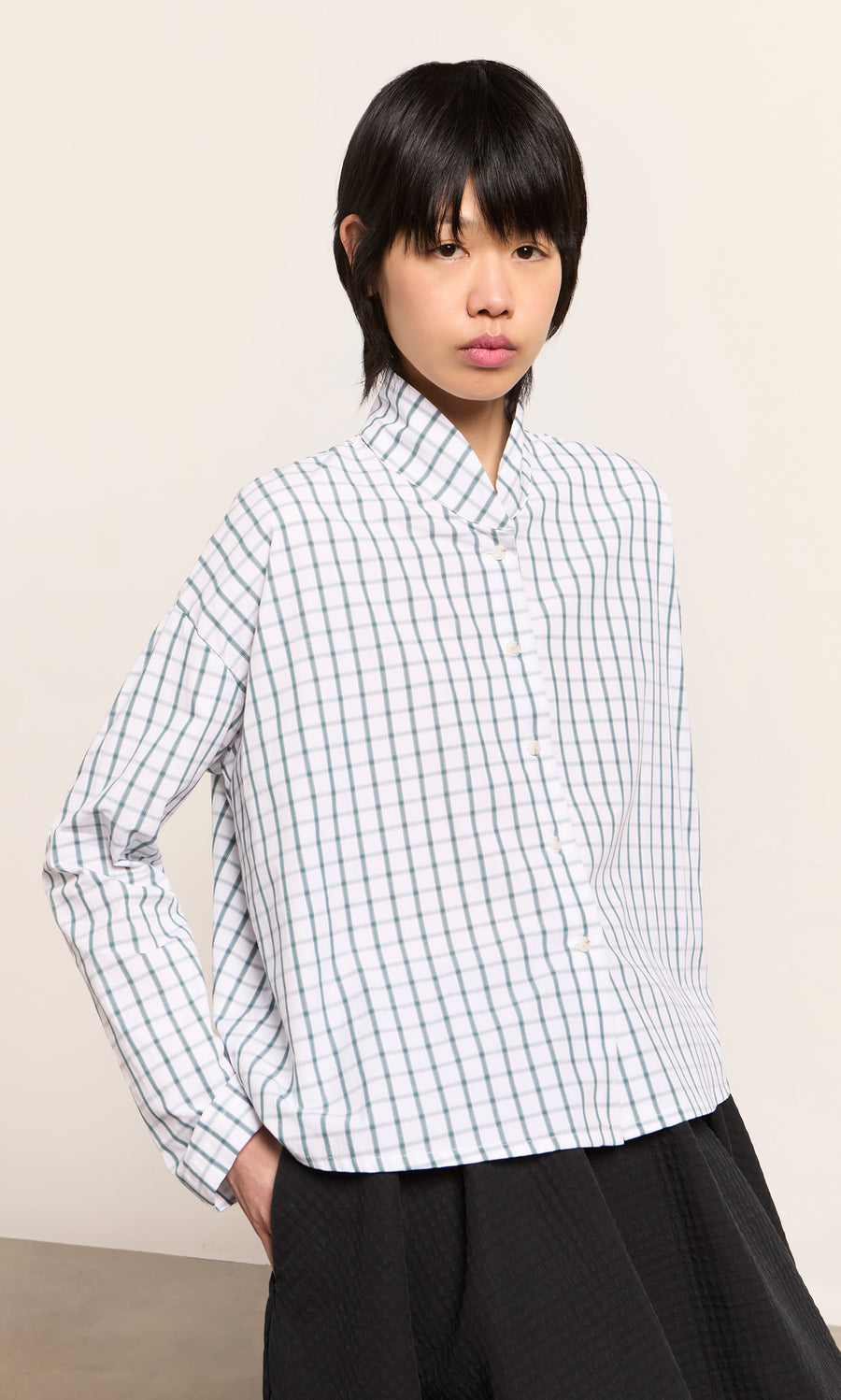 Plümo  Person in the green grid check Bruges Shirt and black pants stands hands-in-pockets against a plain background.