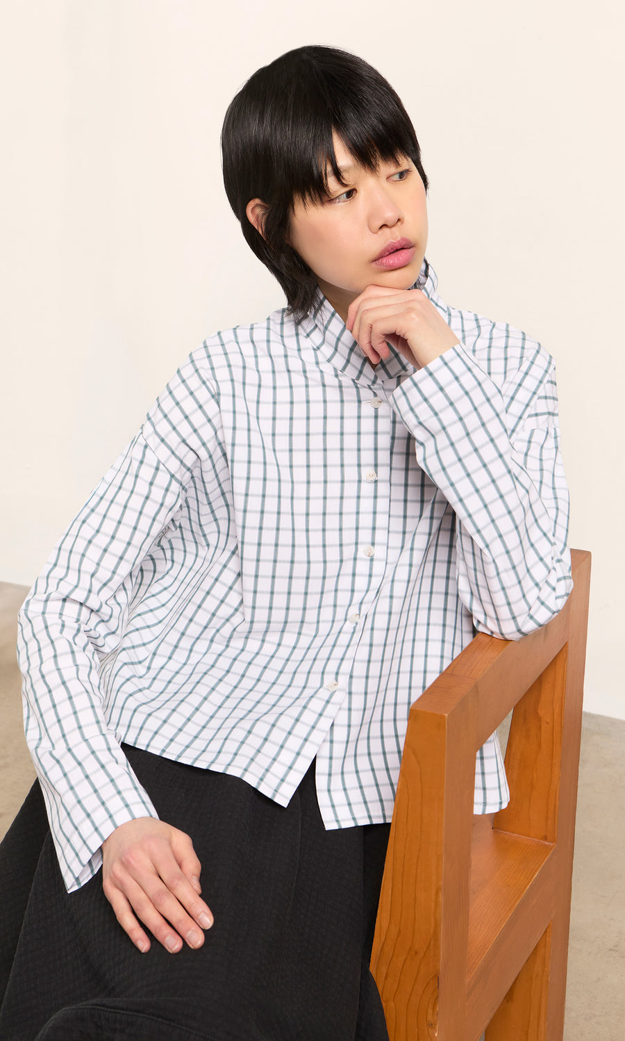 Plümo  Person with short black hair in a Bruges Shirt sits on a wooden chair, gazing thoughtfully to the side.