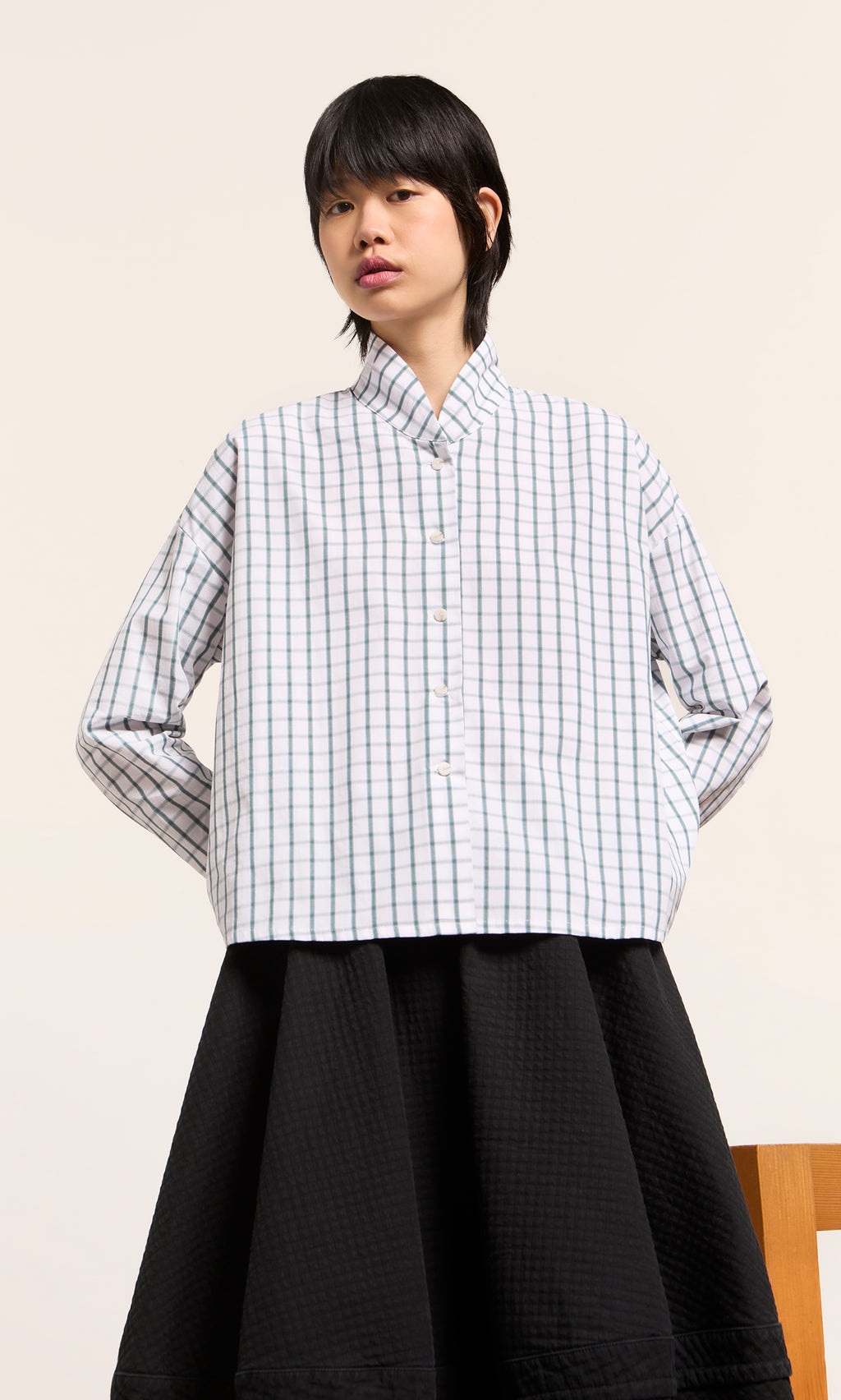 Plümo  Person in a green grid check Bruges Shirt and black quilted skirt, hands behind back. Made in England.