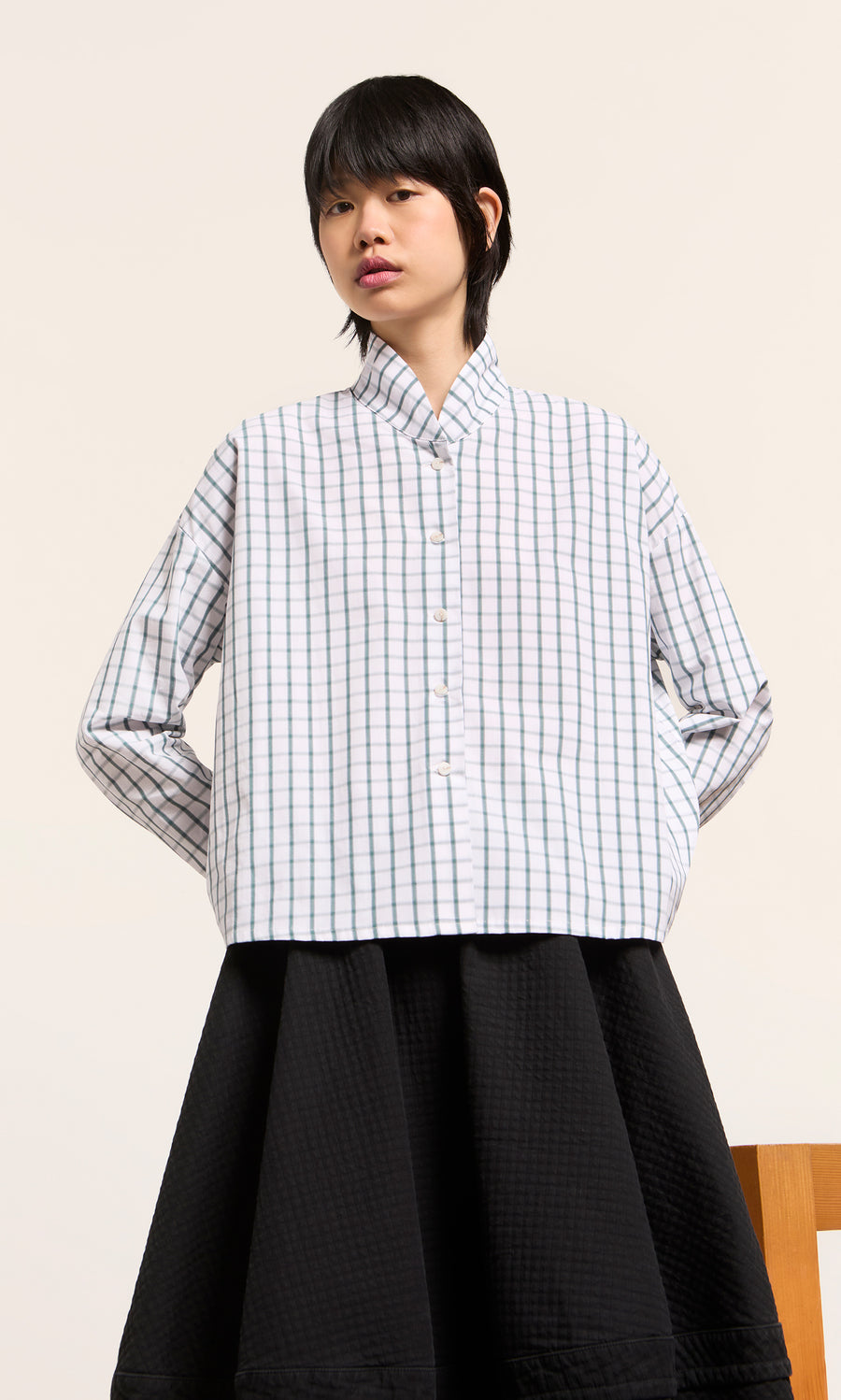 Plümo  Person in a green grid check Bruges Shirt and black quilted skirt, hands behind back. Made in England.