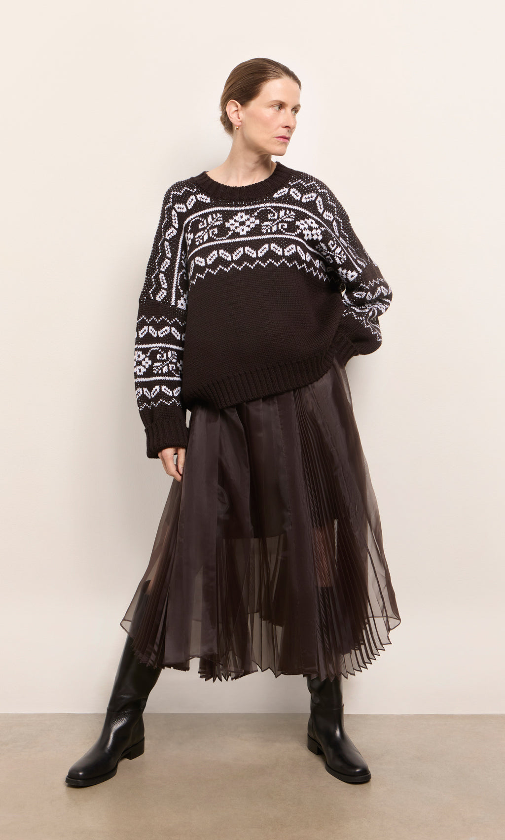 Plümo  Woman in a Kyffin Organza Skirt, black and white sweater, and black boots stands against a plain wall.
