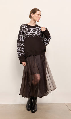 Plümo  Woman in patterned sweater, Kyffin Organza Skirt, and black boots stands against a plain beige wall.
