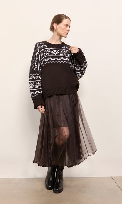 Plümo  Woman in patterned sweater, Kyffin Organza Skirt, and black boots stands against a plain beige wall.
