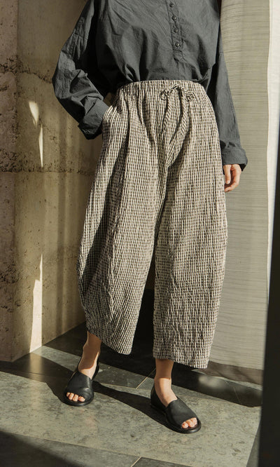 Plümo- Person in a loose black shirt, Butoni Pants - Gingham, and black sandals stands in sunlight by a concrete wall.