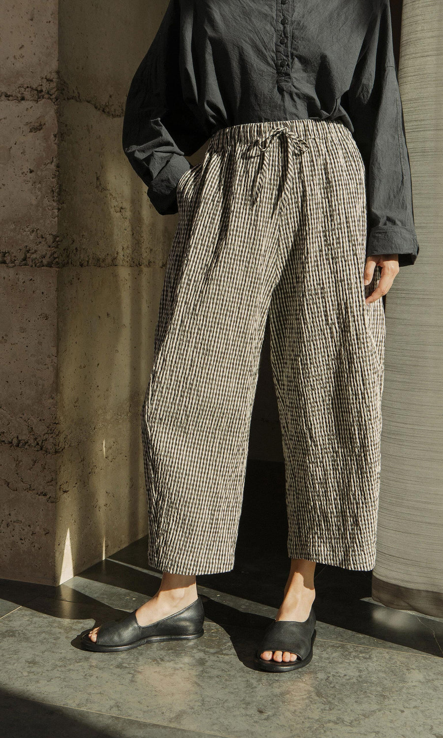 Plümo- Person wearing Butoni Pants - Gingham and black sandals, standing in sunlight with hands in pockets.