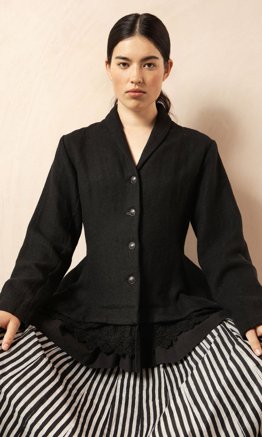 Plümo- Woman with long dark hair in the Cara Riding Jacket and a striped skirt, seated against a light background.