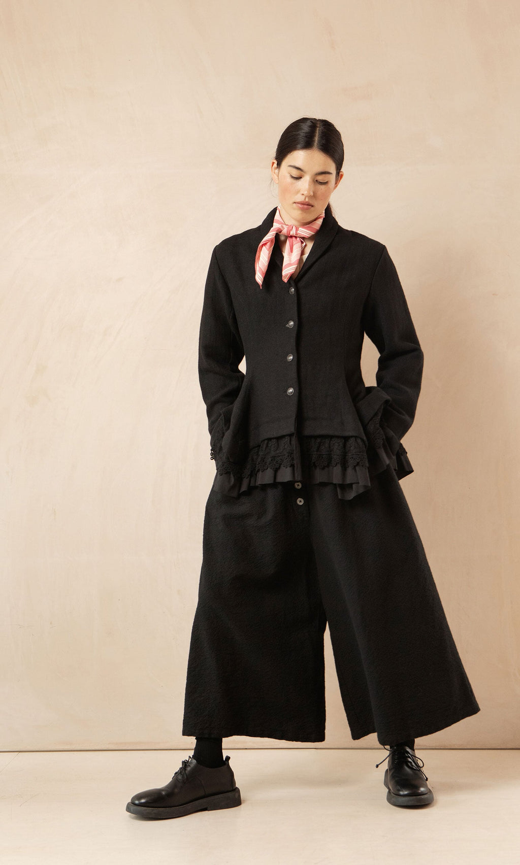 Plümo- Woman in a Cara Riding Jacket and wide-leg pants with a pink scarf, black shoes, stands against a beige background.