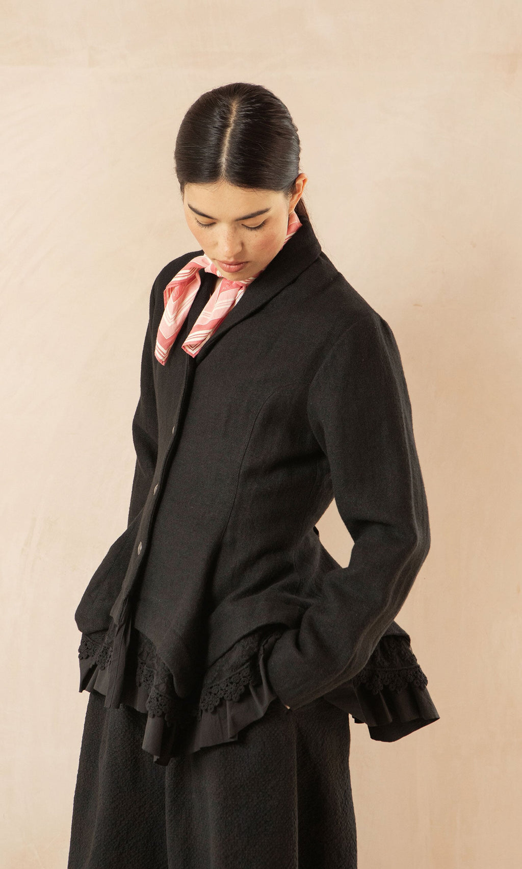 Plümo- Woman in the Cara Riding Jacket with ruffled hem and pink scarf looks down against a beige background.