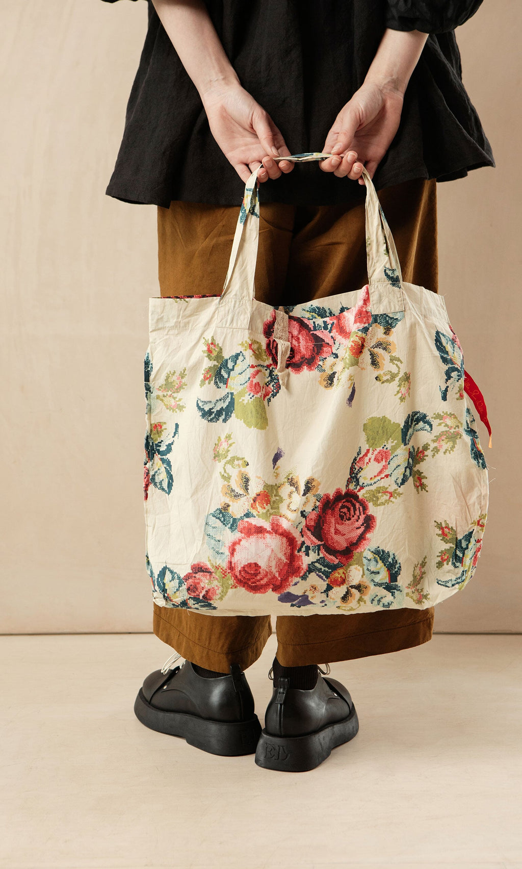 Plümo- Person holding the Carrie Flower Bag, a lightweight cotton tote with floral cross-stitch print, wearing black attire.