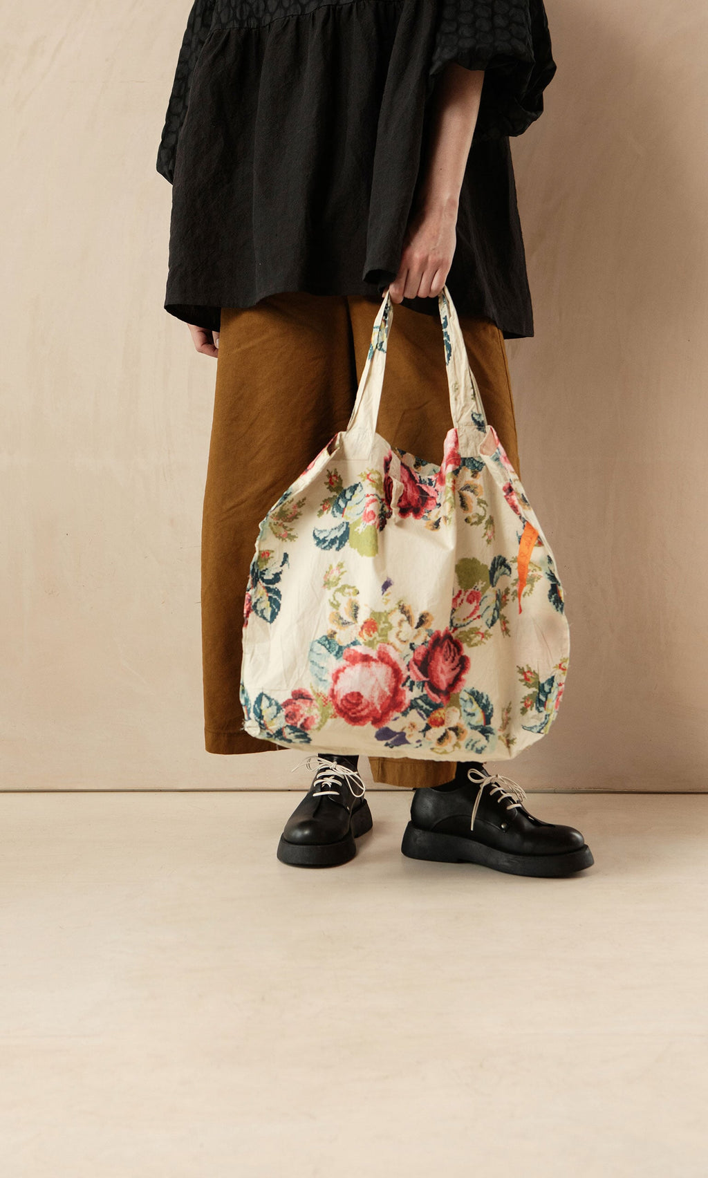Plümo- Person standing, holding a Carrie Flower Bag, wearing brown pants and a black top.