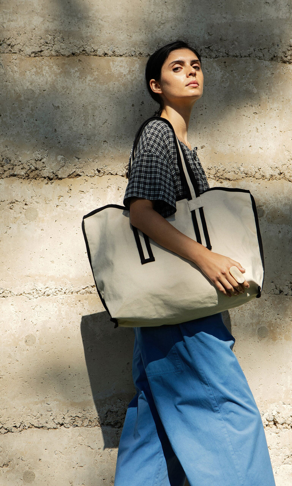 Plümo- Woman in a plaid shirt and blue pants holds the Carryall Bag, standing by a cement wall in sunlight.