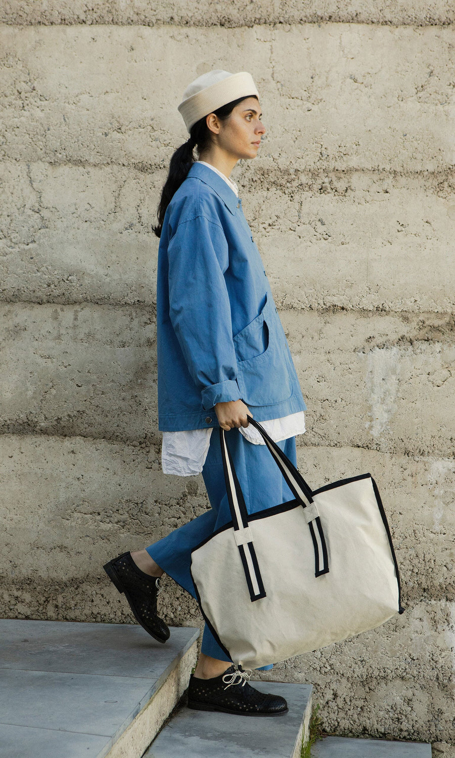 Plümo- Woman in a blue outfit and white hat carries the Carryall Bag with black handles up the stairs.