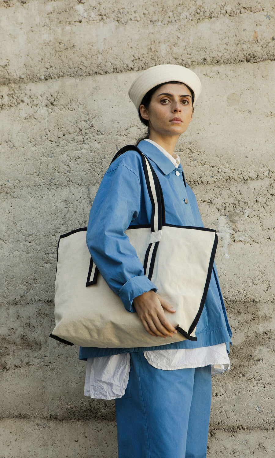 Plümo- Person in blue outfit and white hat holding a Carryall Bag, standing by a textured concrete wall.