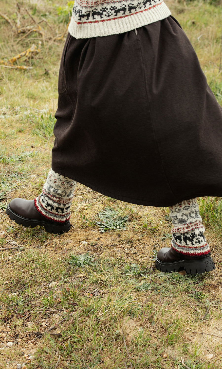 Plümo- Person in a long dark skirt, Cat Tank Top, and black shoes walks on grassy ground.