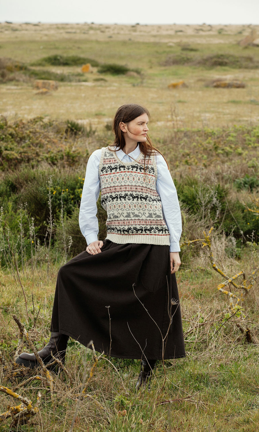 Plümo- A woman in a Cat Tank Top, handknitted in Nepal, and a dark skirt stands in a grassy field, looking to the side.