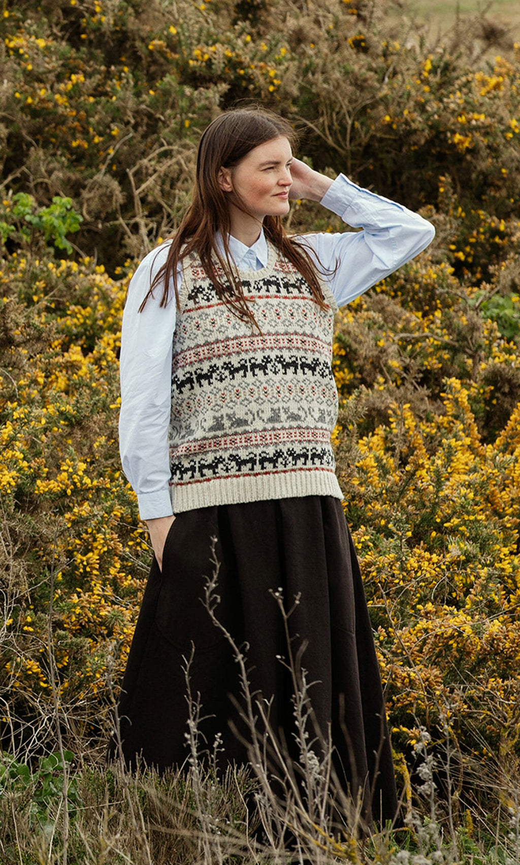 Plümo- Woman stands in a field of yellow gorse, wearing a Cat Tank Top and long dark skirt, handknitted in Nepal.