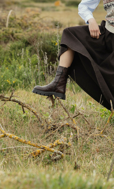 Plümo- A person in a long dark skirt steps over a branch in grassy terrain, wearing Paola Boots with greenery in the background.