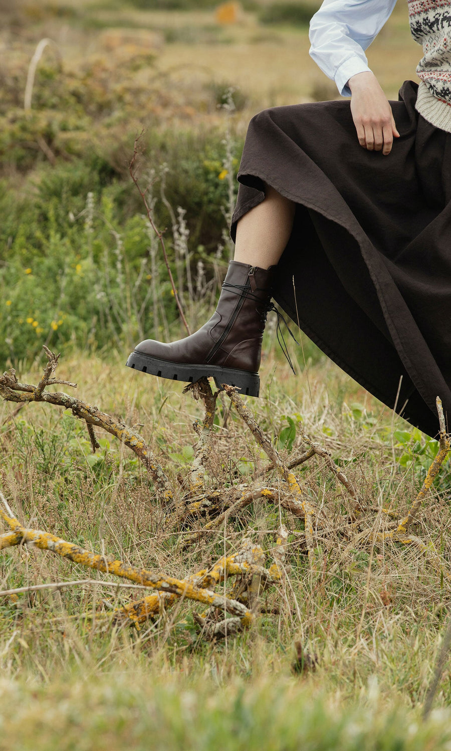 Plümo- A person in a long dark skirt steps over a branch in grassy terrain, wearing Paola Boots with greenery in the background.