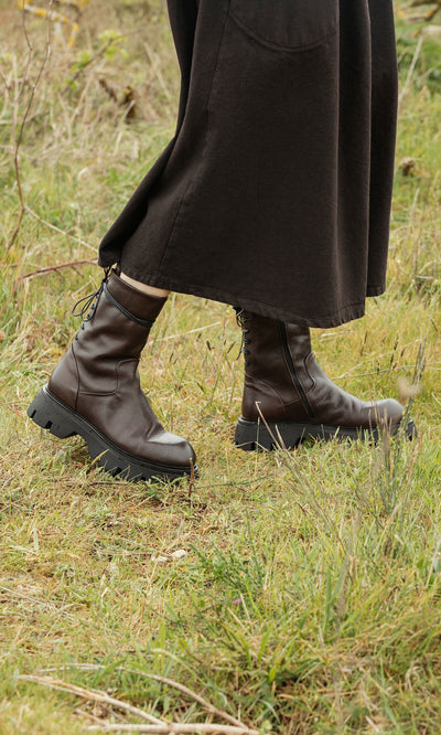 Plümo- A person in a long dark skirt wears Paola Boots while walking on grassy ground outdoors.